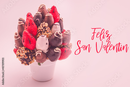 Happy valentine's day greeting card in Spanish that reads Feliz San Valentin with red lettering; A bundle of edible flowers, arrangement of strawberries covered in chocolate isolated on white