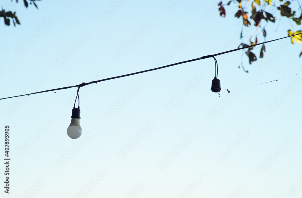 Selecitve focus light bulbs hanging on the electricity wire with blurred leaves and clear sky in background Selecitve focus light bulbs hanging on the electricity wire with blurred leaves and clear sky in background