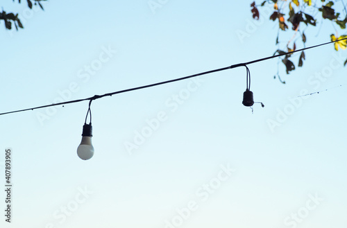 Selecitve focus light bulbs hanging on the electricity wire with blurred leaves and clear sky in background