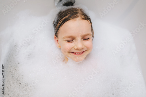 A small, smiling, cute red-haired with long hair, a child bathes, lying in a white bath with foam from soap and shampoo. 
