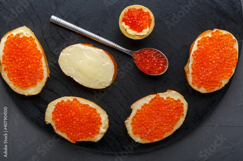 Canapes with butter and red caviar and a tartlet and a spoon with caviar on a black board on a concrete background.