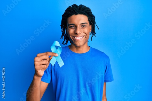 Young african american man holding blue ribbon looking positive and happy standing and smiling with a confident smile showing teeth photo