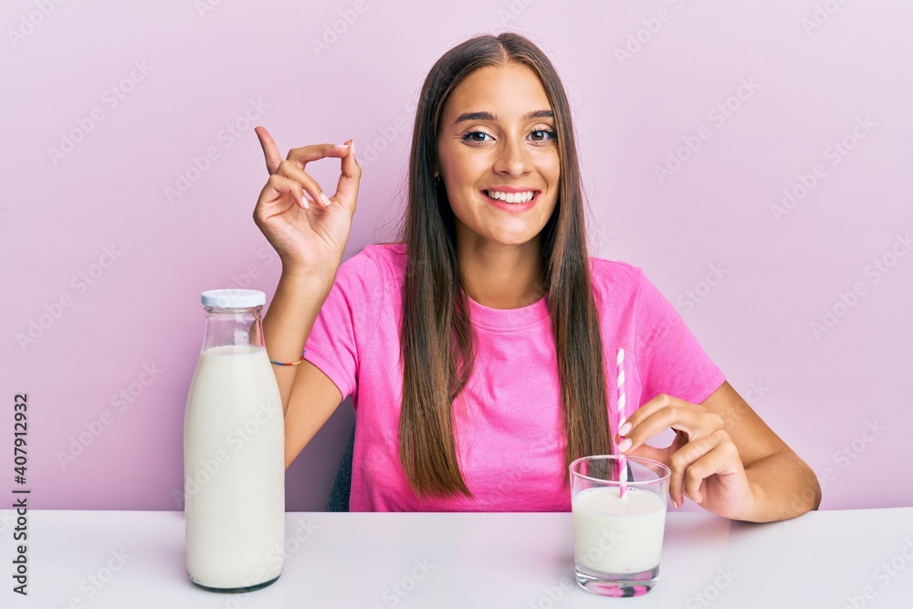 Young hispanic woman drinking a glass of fresh milk sitting on the table smiling happy pointing with hand and finger to the side Young hispanic woman drinking a glass of fresh milk sitting on the table smiling happy pointing with hand and finger to the side