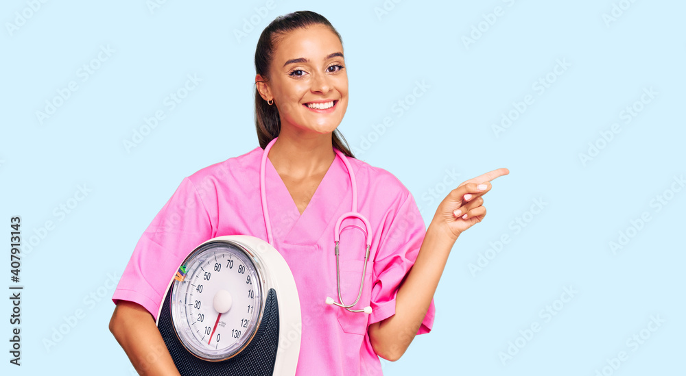Young hispanic woman as nutritionist doctor holding weighing machine smiling happy pointing with hand and finger to the side Young hispanic woman as nutritionist doctor holding weighing machine smiling happy pointing with hand and finger to the side