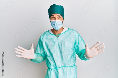 Young handsome man wearing surgeon uniform and medical mask crazy and mad shouting and yelling with aggressive expression and arms raised. frustration concept.