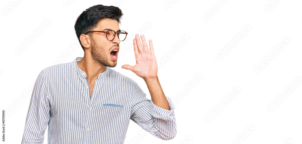 Young handsome man wearing casual clothes and glasses shouting and screaming loud to side with hand on mouth. communication concept. Young handsome man wearing casual clothes and glasses shouting and screaming loud to side with hand on mouth. communication concept.