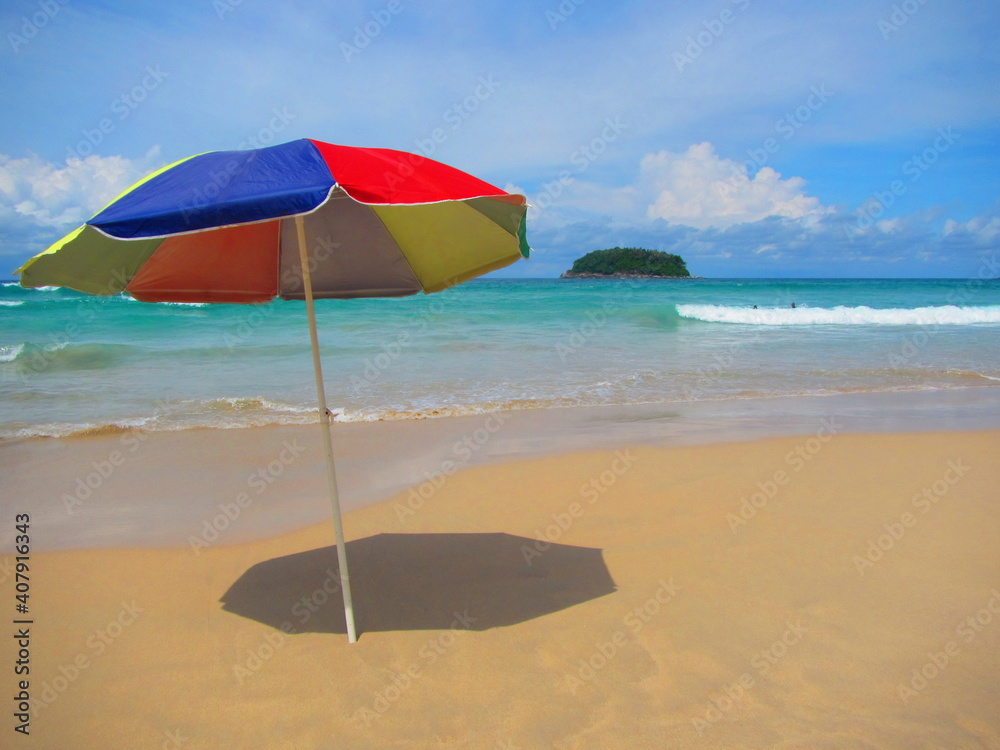 Phuket, Thailand, October 02, 2018: An isolated multi-coloured umbrella flutters in the wind on a golden beach and aquamarine blue water with a lone tree capped island in the background.