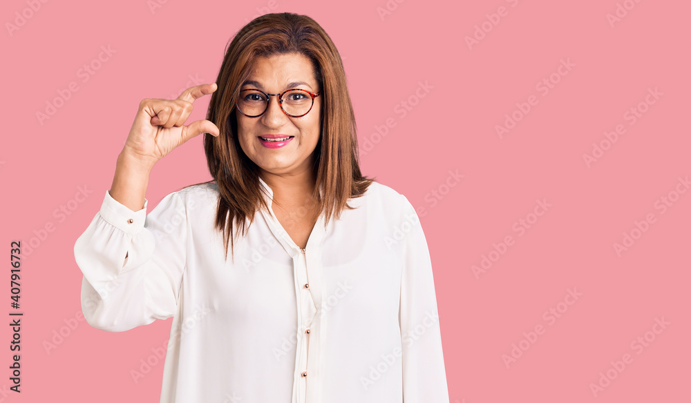 Middle age latin woman wearing casual clothes and glasses smiling and confident gesturing with hand doing small size sign with fingers looking and the camera. measure concept.