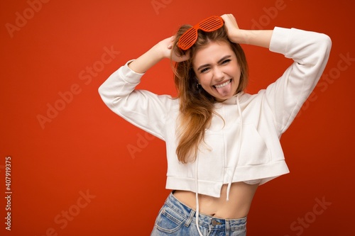 Shot of attractive funny smiling young blonde woman isolated over orange background wearing casual white hoodie and stylish sunglasses enjoying and having fun