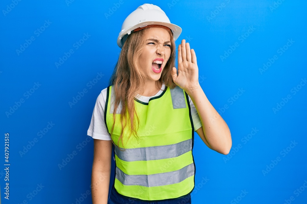Beautiful blonde caucasian woman wearing architect hardhat shouting and screaming loud to side with hand on mouth. communication concept. Beautiful blonde caucasian woman wearing architect hardhat shouting and screaming loud to side with hand on mouth. communication concept.