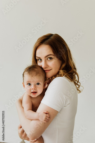 Beautiful mom hugs and kisses the newborn in the studio on a white background. A young mother in a white bodysuit hugs and kisses her newborn daughter in the studio.