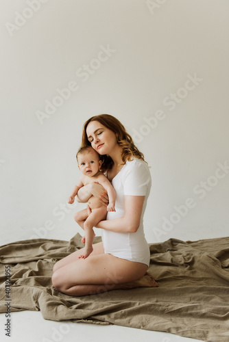 Beautiful mom hugs and kisses the newborn in the studio on a white background. A young mother in a white bodysuit hugs and kisses her newborn daughter in the studio.