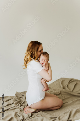 Beautiful mom hugs and kisses the newborn in the studio on a white background. A young mother in a white bodysuit hugs and kisses her newborn daughter in the studio.