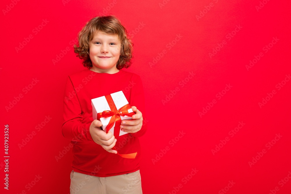 Fototapeta premium Photo shot of joyful smiling blonde curly boy isolated over red background wall wearing red sweater holding gift box and looking at camera