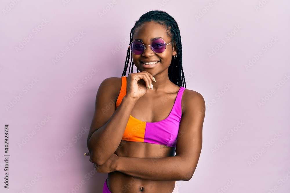 Young african american woman wearing bikini and sunglasses looking confident at the camera with smile with crossed arms and hand raised on chin. thinking positive.
