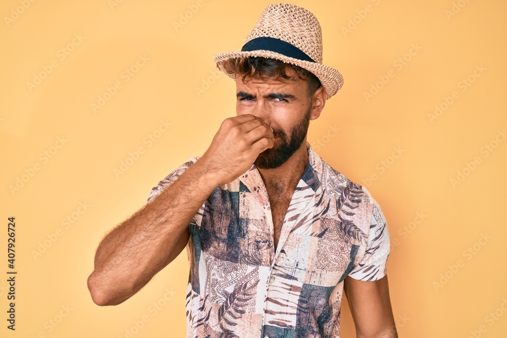 Young hispanic man wearing summer hat smelling something stinky and disgusting, intolerable smell, holding breath with fingers on nose. bad smell Young hispanic man wearing summer hat smelling something stinky and disgusting, intolerable smell, holding breath with fingers on nose. bad smell