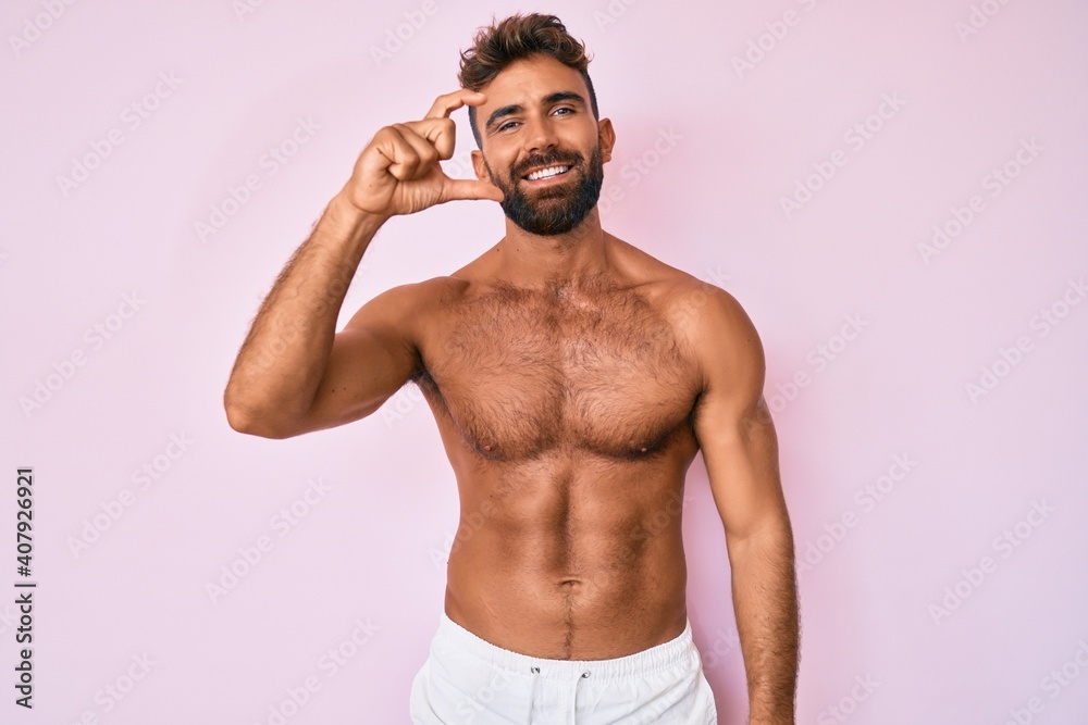 Fototapeta premium Young hispanic man standing shirtless smiling and confident gesturing with hand doing small size sign with fingers looking and the camera. measure concept.