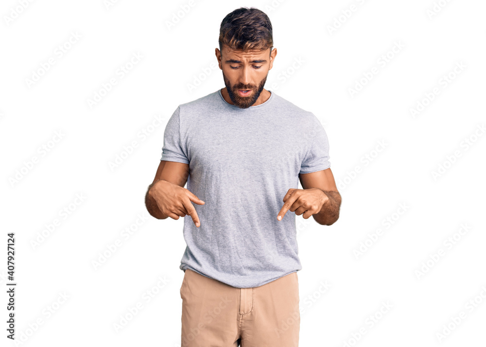 Young hispanic man wearing casual clothes pointing down with fingers showing advertisement, surprised face and open mouth Young hispanic man wearing casual clothes pointing down with fingers showing advertisement, surprised face and open mouth