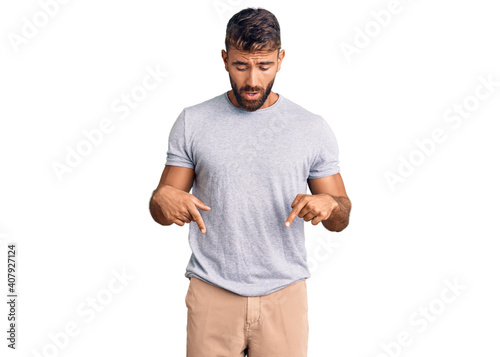 Young hispanic man wearing casual clothes pointing down with fingers showing advertisement, surprised face and open mouth