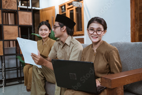 Asian woman teacher in civil servant uniform smiling looks at the camera holding a laptop computer while working with the team at home photo