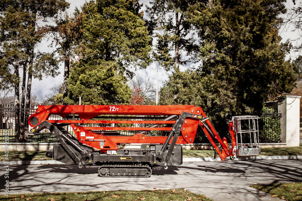 Red remote control tracked lift designed for tree work with basket parked in front of estate fence with evergreen trees Red remote control tracked lift designed for tree work with basket parked in front of estate fence with evergreen trees
