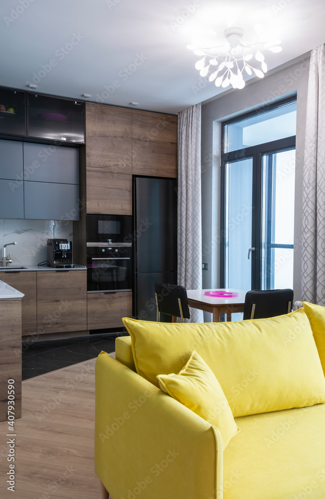 Fototapeta premium The interior of the studio kitchen is decorated in gray tones with wooden veneers. Kitchen appliances are black. In the foreground is a yellow sofa. Vertical.