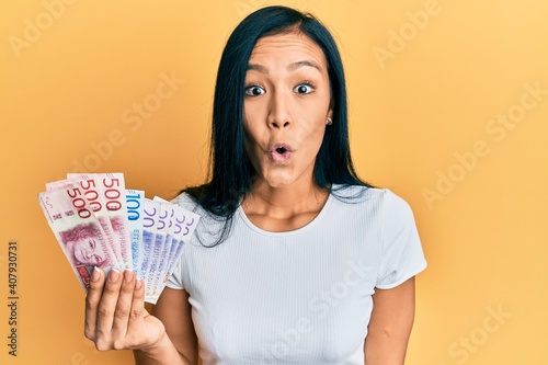 Beautiful hispanic woman holding swedish krona banknotes scared and amazed with open mouth for surprise, disbelief face