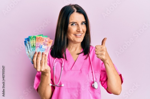 Middle age brunette doctor woman holding swiss francs smiling happy and positive, thumb up doing excellent and approval sign