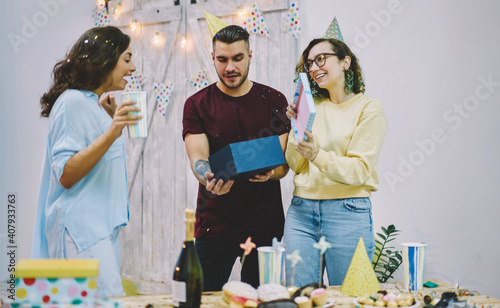 Young hipsters greeting girl on her birthday party opening decorative box with surprise during celebration, happy young woman amazed with gift from best friends satisfied with holiday and festive mood