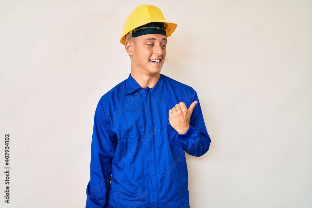 Young hispanic boy wearing worker uniform and hardhat smiling with happy face looking and pointing to the side with thumb up.