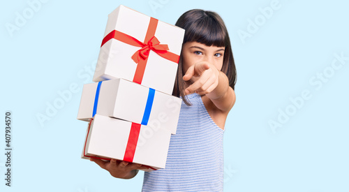 Young little girl with bang holding gifts pointing with finger to the camera and to you, confident gesture looking serious