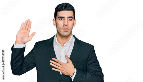 Obraz na plátně Handsome hispanic man wearing business clothes swearing with hand on chest and o