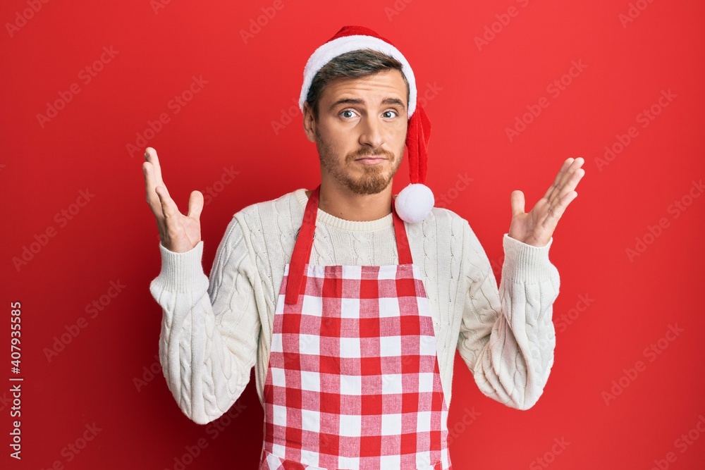 Naklejka premium Handsome caucasian man wearing baker apron and christmas hat clueless and confused with open arms, no idea and doubtful face.