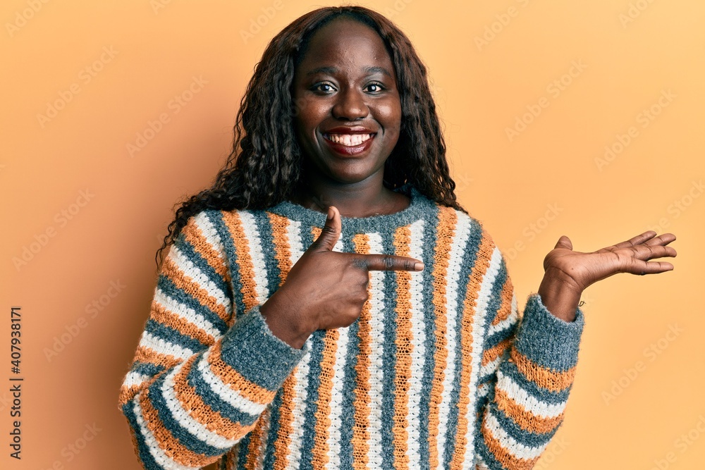 African young woman wearing wool winter sweater amazed and smiling to the camera while presenting with hand and pointing with finger. African young woman wearing wool winter sweater amazed and smiling to the camera while presenting with hand and pointing with finger.