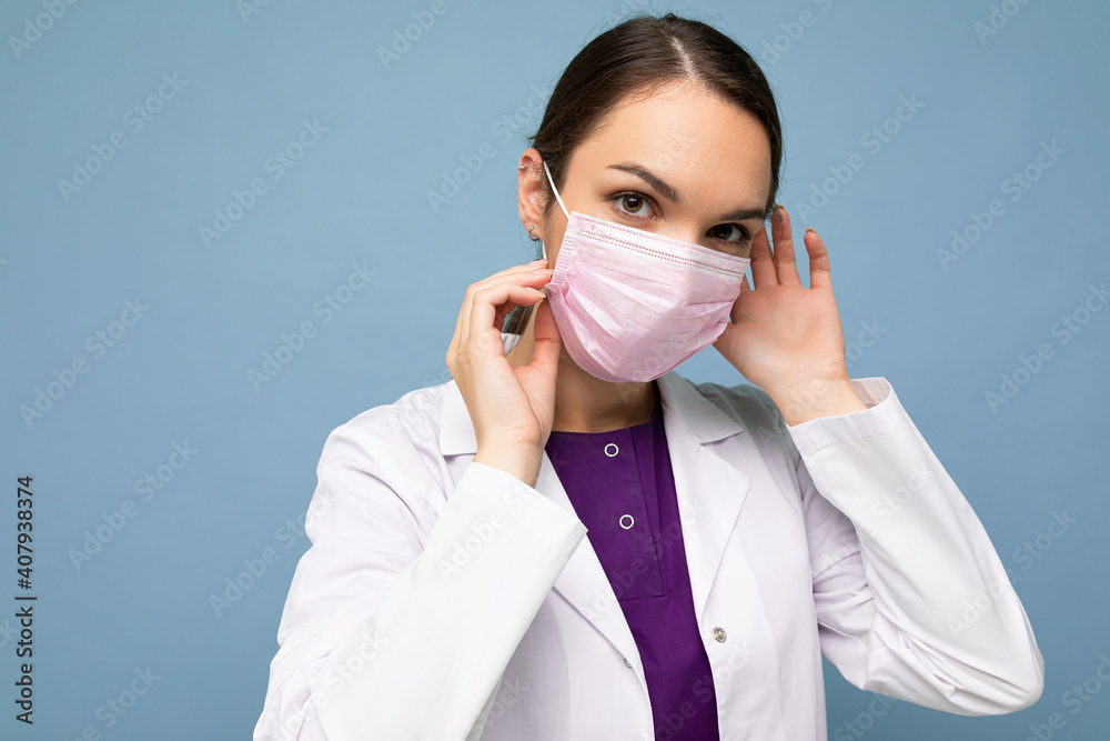 Portrait of an attractive young female doctor in white coat and medical mask standing isolated on blue background Portrait of an attractive young female doctor in white coat and medical mask standing isolated on blue background