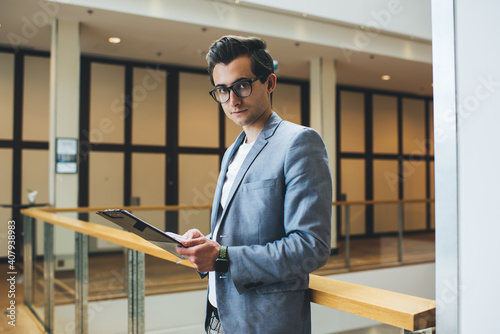 Half length portrait of handsome serious male manager in eyewear holding planner for organizing job process, serious caucasian businessman holding finance documents looking at camera in modern office