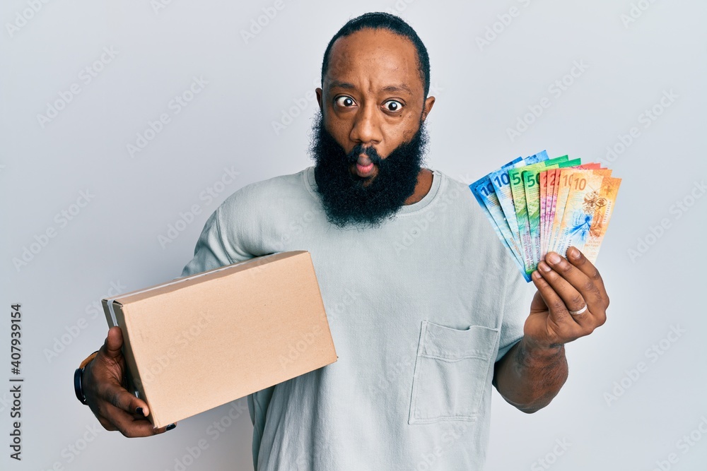 Young african american man holding delivery box and swiss franc banknotes making fish face with mouth and squinting eyes, crazy and comical. Young african american man holding delivery box and swiss franc banknotes making fish face with mouth and squinting eyes, crazy and comical.