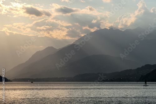 A man stand up paddling during the sunset on Millstaetter lake in Austria. The lake is surrounded by high Alps. Calm surface of the lake reflecting the sunbeams. The sun sets behind the mountains.