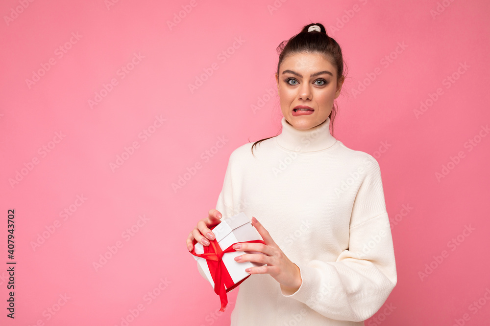 Attractive amazed upset young brunette female person isolated over pink background wall wearing white sweater holding gift box and unboxing present looking at camera Attractive amazed upset young brunette female person isolated over pink background wall wearing white sweater holding gift box and unboxing present looking at camera