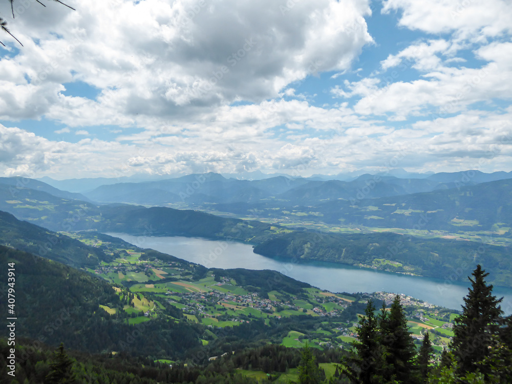 A panoramic view on the Millstaettersee lake from an Alpine pasture in Austria. The distant lake is surrounded by high mountains. Few clouds above. Endless mountain chains. Lush and vast forest. Relax