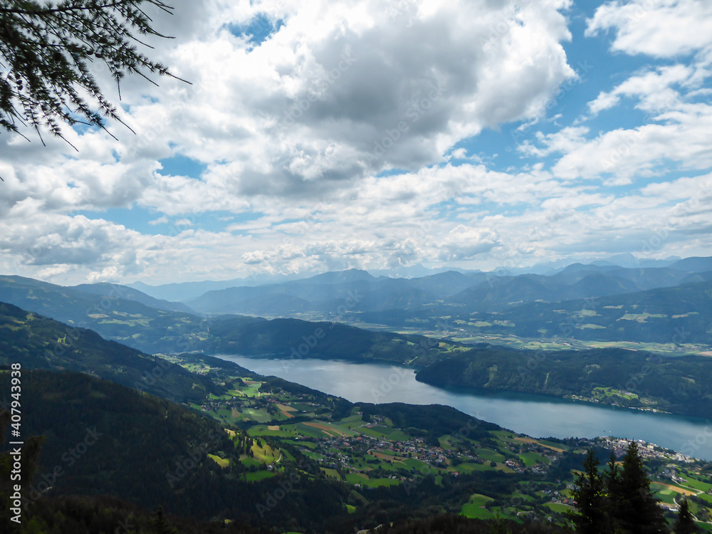A panoramic view on the Millstaettersee lake from an Alpine pasture in Austria. The distant lake is surrounded by high mountains. Few clouds above. Endless mountain chains. Lush and vast forest. Relax