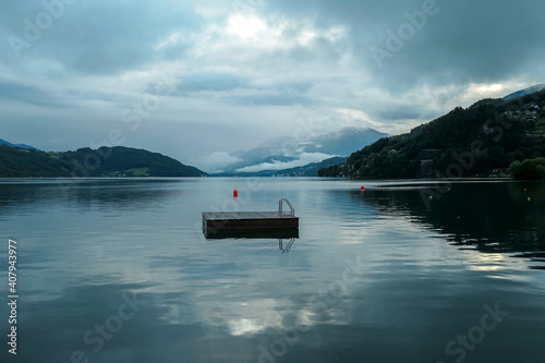 A panoramic view on the Millstaettersee lake from its shore. The lake is surrounded by high mountains. A wooden platform drifting on its surface. Thick and dark clouds above. Serenity and calmness photo