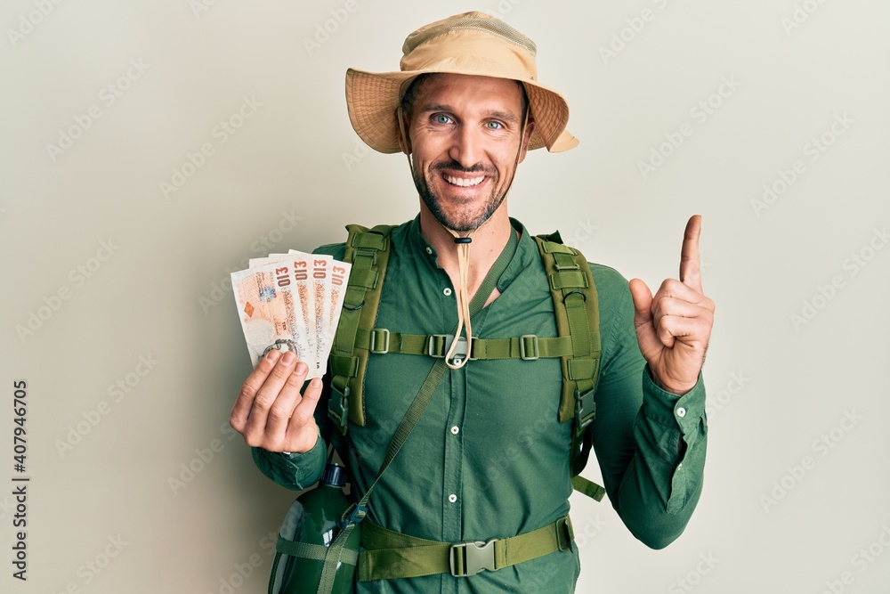 Handsome man with beard wearing explorer hat holding 10 pounds smiling happy pointing with hand and finger to the side Handsome man with beard wearing explorer hat holding 10 pounds smiling happy pointing with hand and finger to the side