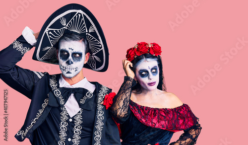 Young couple wearing mexican day of the dead costume over background confuse and wondering about question. uncertain with doubt, thinking with hand on head. pensive concept.