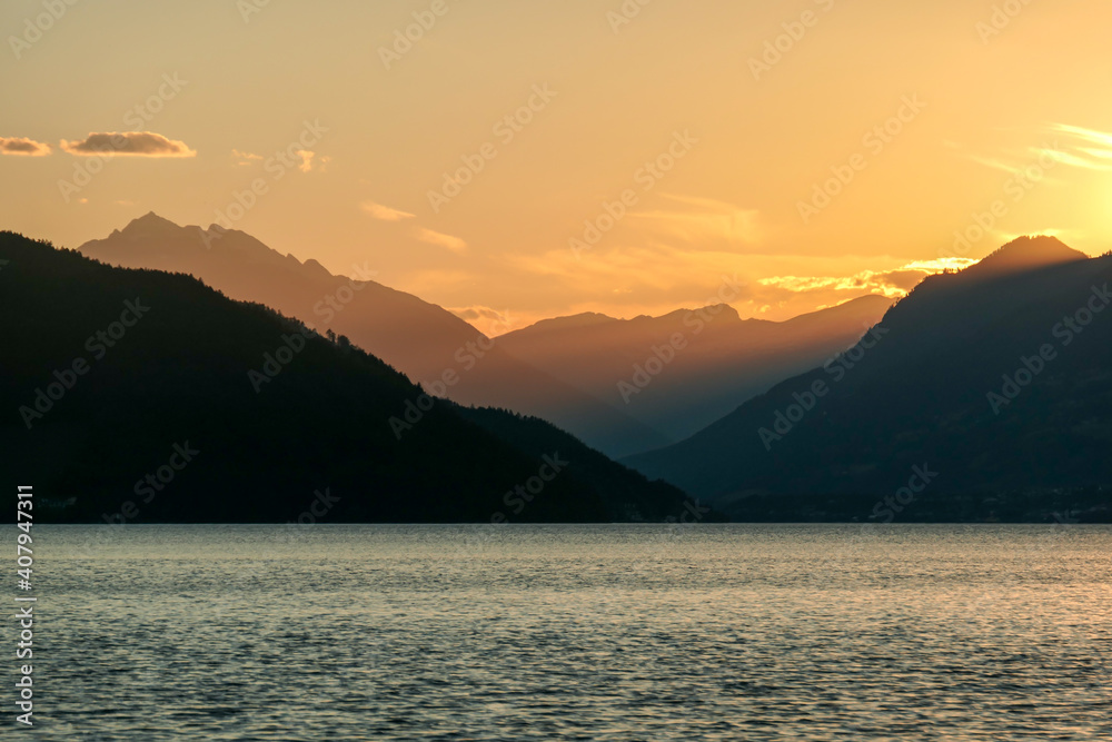 A sunset by Millstaetter lake in Austria. The lake is surrounded by high Alps. Calm surface of the lake reflecting the sunbeams. The sun sets behind the mountains. A bit of overcast. Natural beauty