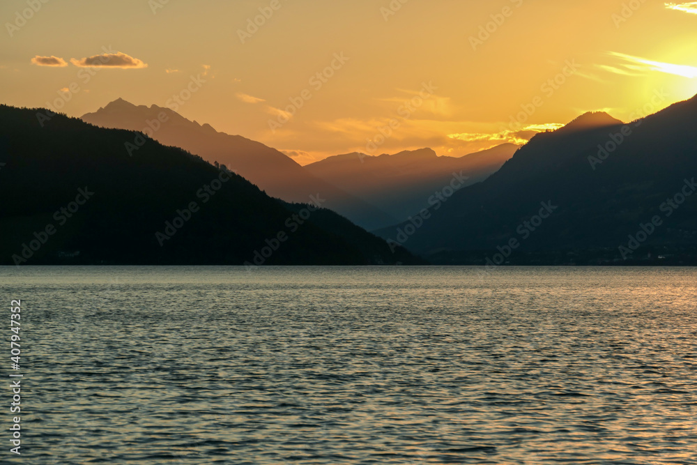 A sunset by Millstaetter lake in Austria. The lake is surrounded by high Alps. Calm surface of the lake reflecting the sunbeams. The sun sets behind the mountains. A bit of overcast. Natural beauty