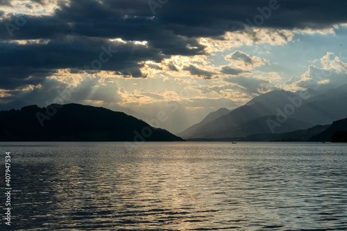 A sunset by Millstaetter lake in Austria. The lake is surrounded by high Alps. Calm surface of the lake reflecting the sunbeams. The sun sets behind the mountains. Thick clouds above. Natural beauty