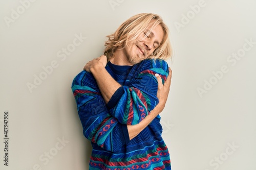 Caucasian young man with long hair wearing colorful winter sweater hugging oneself happy and positive, smiling confident. self love and self care