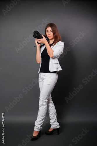 A woman in a white suit with a camera in her hands posing while standing on a black background. Contemporary photographer in the studio.