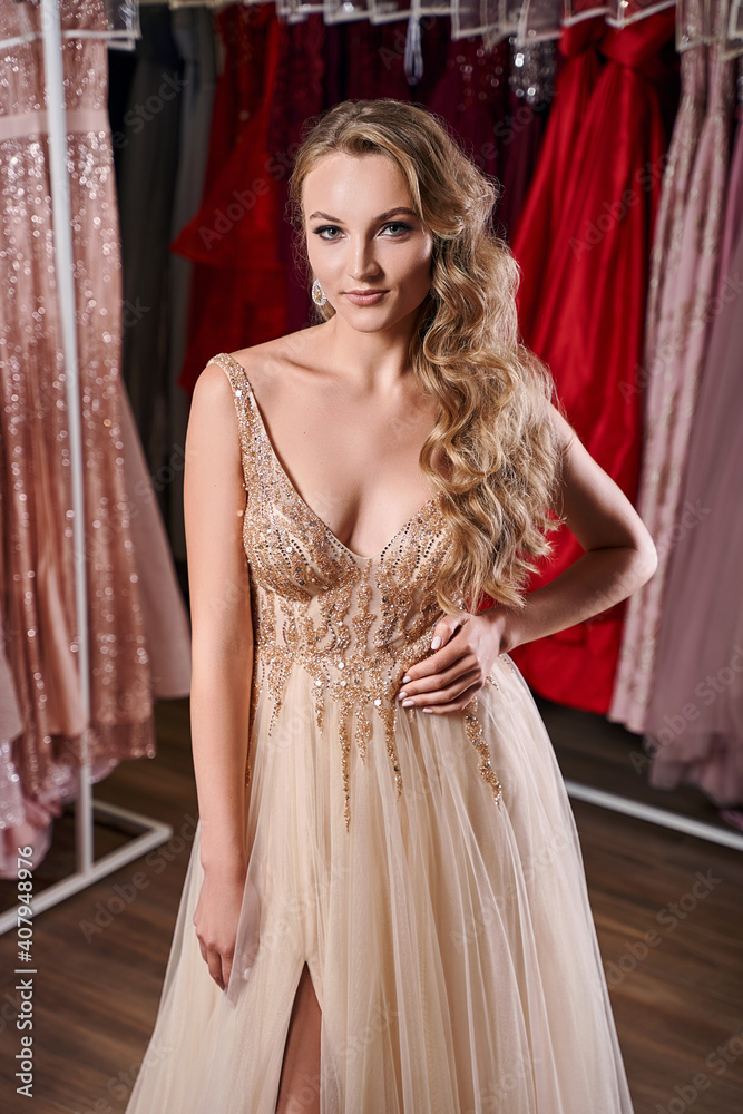 Beautiful blonde girl wearing a full-length yellow champaign chiffon slit prom ball gown decorated with golden sequins. Model in dress hire service with many dresses in different color and style.